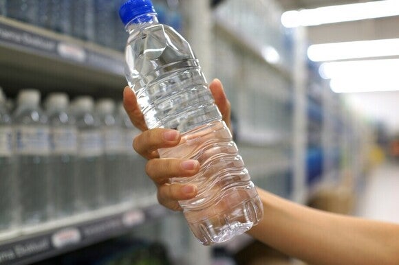 Person hält eine volle Flasche Wasser in der Hand mit einem blauen Deckel und vor einer Regalwand voller Wasserflaschen.