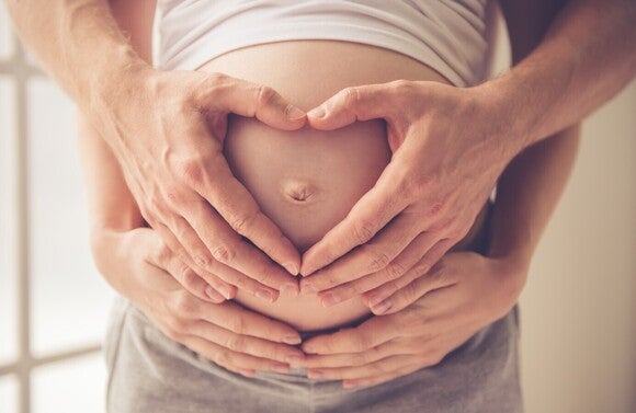 Zwei Paar Hände formen gemeinsam ein Herz auf einem schwangeren Bauch.