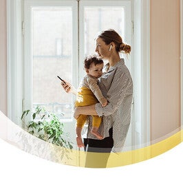 Frau vor Fenster mit ihrem Baby auf dem Arm und aufs Handy schauend